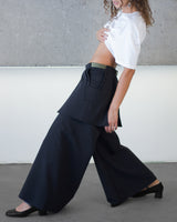 Person wearing a white blouse and black wide-leg pants against a gray wall.