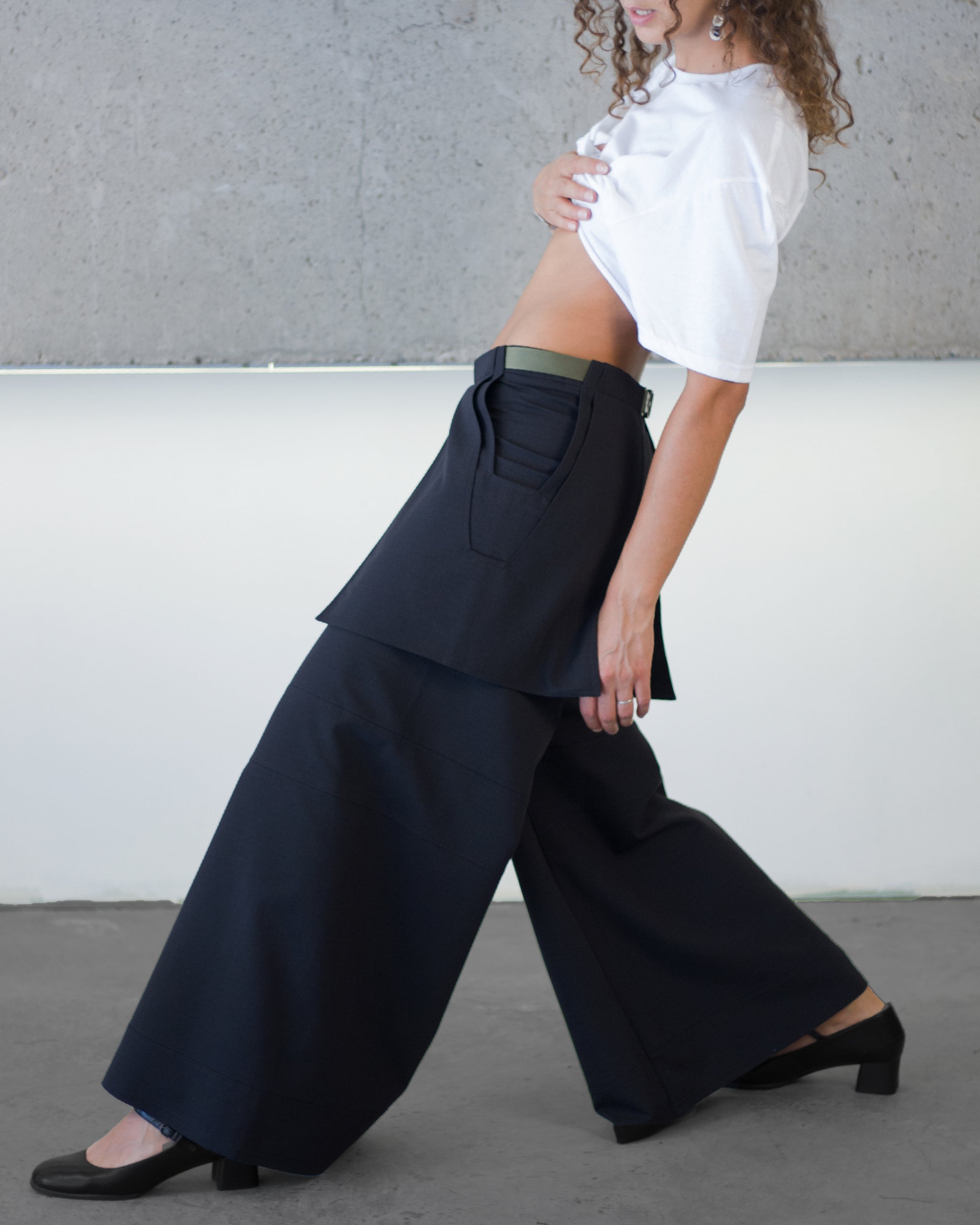 Person wearing a white blouse and black wide-leg pants against a gray wall.
